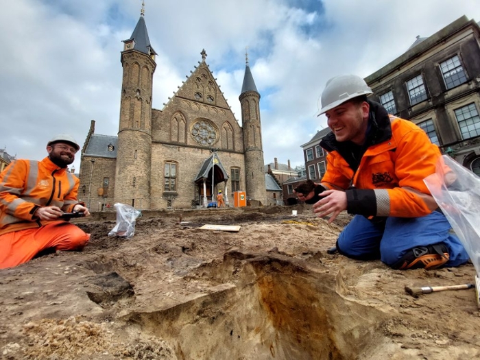 Twee mannen in oranje werkkleding en met helm op zitten op hun knieën in het zand voor de Ridderzaal.