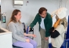 Een vrouw zit op een stoel, terwijl een man haar arm onderzoekt met een meetinstrument, en een meisje met een witte gans-hoed toekijkt.