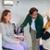 Een vrouw zit op een stoel, terwijl een man haar arm onderzoekt met een meetinstrument, en een meisje met een witte gans-hoed toekijkt.
