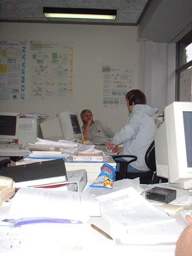 Voordat pdf’s de norm werden, bestond onderzoek vooral op papier. Op deze foto uit 2001 bespreken promovendi Todor Stefanov en Vladimir Zivkovic onderzoeksideeën in hun kantoor (kamer 122, Snellius), omringd door grote computers, onderzoeksposters en stapels geprinte artikelen en rapporten.
